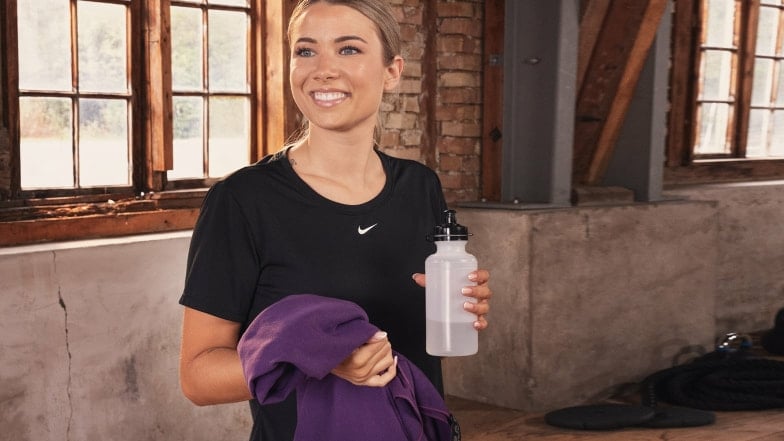 Eine lächelnde Frau in Sportkleidung hält eine Wasserflasche und Handtuch.