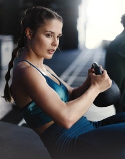 Frau sitzt auf einer Matte im Fitnessstudio und hält eine Kettlebell mit beiden Händen vor dem Oberkörper. Die Übung stärkt die Bauch- und Armmuskulatur.