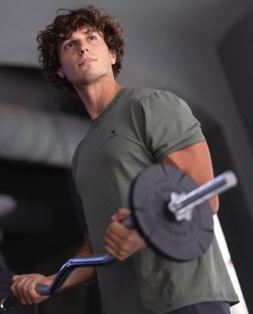 Mann in grünem T-Shirt hebt eine Hantelstange im Fitnessstudio. Blick nach oben gerichtet.