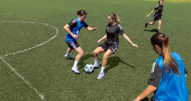 Mehrere Fußballspielerinnen stehen auf einem Kunstrasenplatz im Zweikampf um den Ball. Zu sehen sind Spielerinnen in Trainingskleidung während einer Spielsituation.