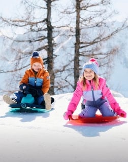 Zwei Kinder in Winterkleidung sitzen lachend auf Schlitten im Schnee.