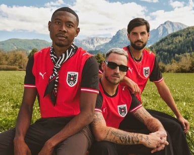 Drei Männer in rot-schwarzen Fußballtrikots sitzen auf einer Wiese vor Berglandschaft. Sie blicken zur Kamera, einer trägt eine Sonnenbrille.