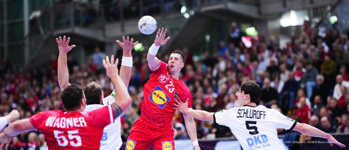 Handballspieler beim Sprungwurf, verteidigt von Gegenspielern vor vollen Zuschauerrängen.