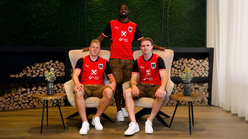 Drei Fußballspieler in roten ÖFB-Trikots und ÖFB-Lederhosen sitzen und stehen vor gestapeltem Brennholz.