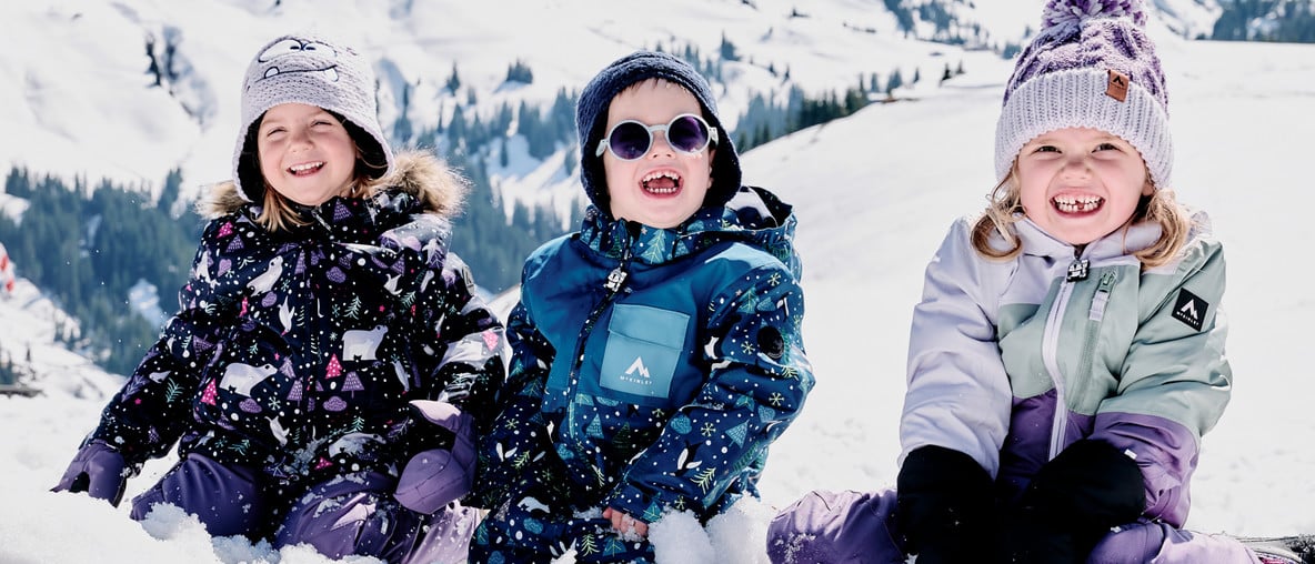 Drei lachende Kinder in Winterkleidung sitzen im Schnee vor einer verschneiten Berglandschaft