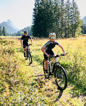 Zwei Mountainbiker fahren auf einem schmalen Weg durch eine Wiese vor bewaldeten Bergen