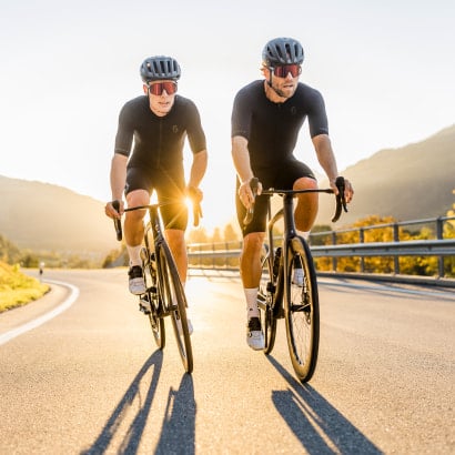 Zwei Radfahrer auf Rennrädern bei Sonnenuntergang auf einer Bergstraße.