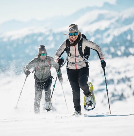 Zwei Personen wandern mit Schneeschuhen und Stöcken durch verschneite Berglandschaft bei Sonnenschein.