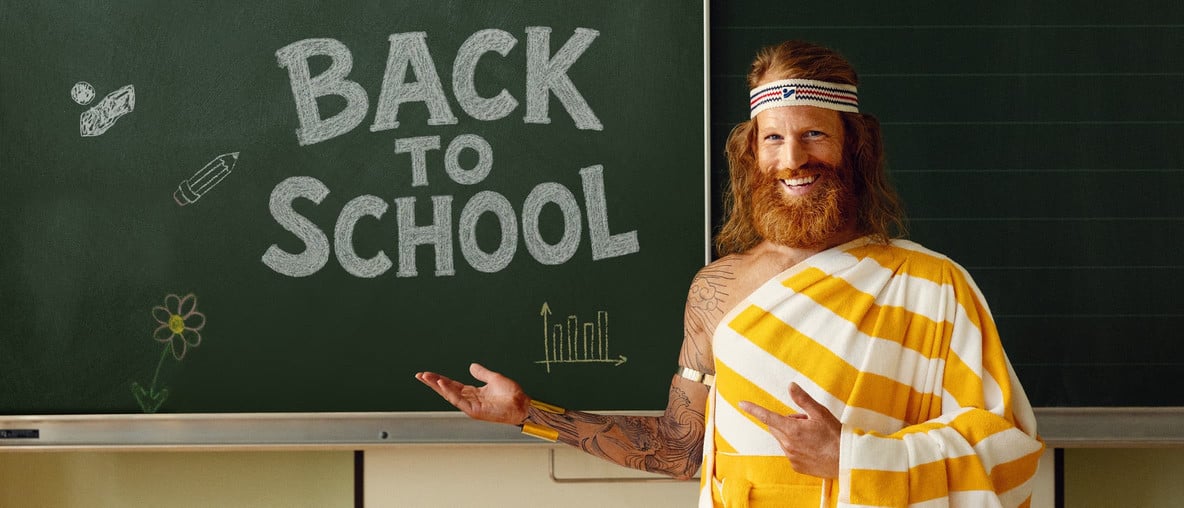 Der Sommer steht vor einer dunkelgrünen Schultafel, auf der mit weißer Kreide "Back To School" geschrieben steht, und lächelt fröhlich.