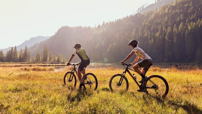 Zwei Personen fahren mit Fahrrädern durch eine Wiese vor Bergen und einem Gewässer.