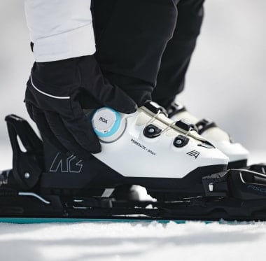 Eine Person in Handschuhen stellt einen weißen Skischuh von K2 mit Boa Verschluss im Schnee ein.