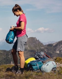 Frau steht auf einem Berg und packt einen Schlafsack neben mehreren Rucksäcken aus.