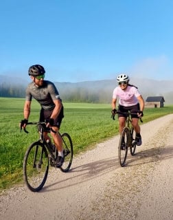Zwei Erwachsene fahren mit Rennrädern auf einem Schotterweg durch eine grüne Landschaft mit Bergen im Hintergrund.