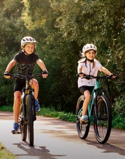 Zwei Kinder fahren mit Helmen Fahrrad auf einem sonnigen Weg.