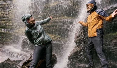 Muž a žena v nepromokavých bundách se smějí pod vodopádem.