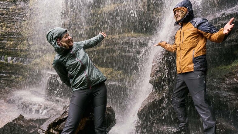 Muž a žena v nepromokavých bundách se smějí pod vodopádem.