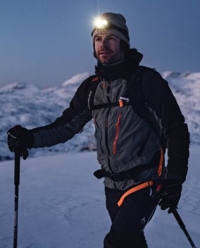 Eine Person mit Stirnlampe und Wanderstöcken steht in winterlicher Berglandschaft bei Dämmerung.