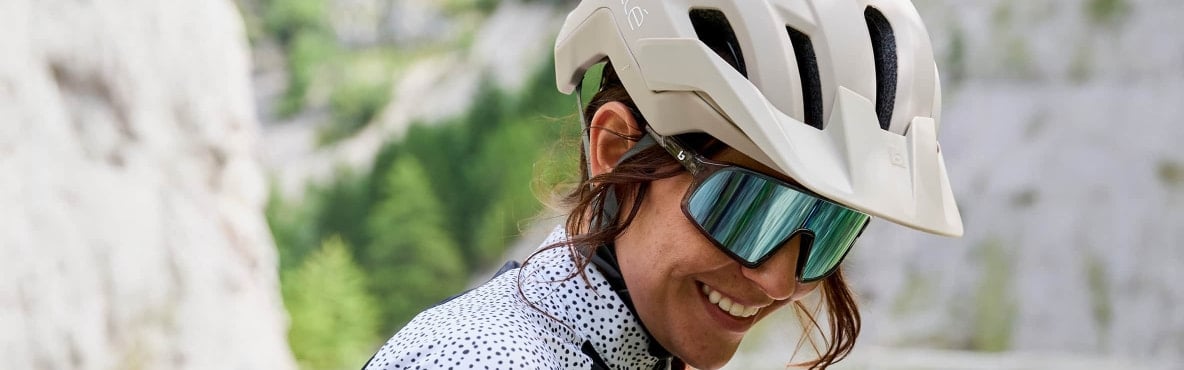 Person mit Fahrradhelm und verspiegelter Sportbrille im Freien. Unscharfe Felsen und Grünflächen im Hintergrund.