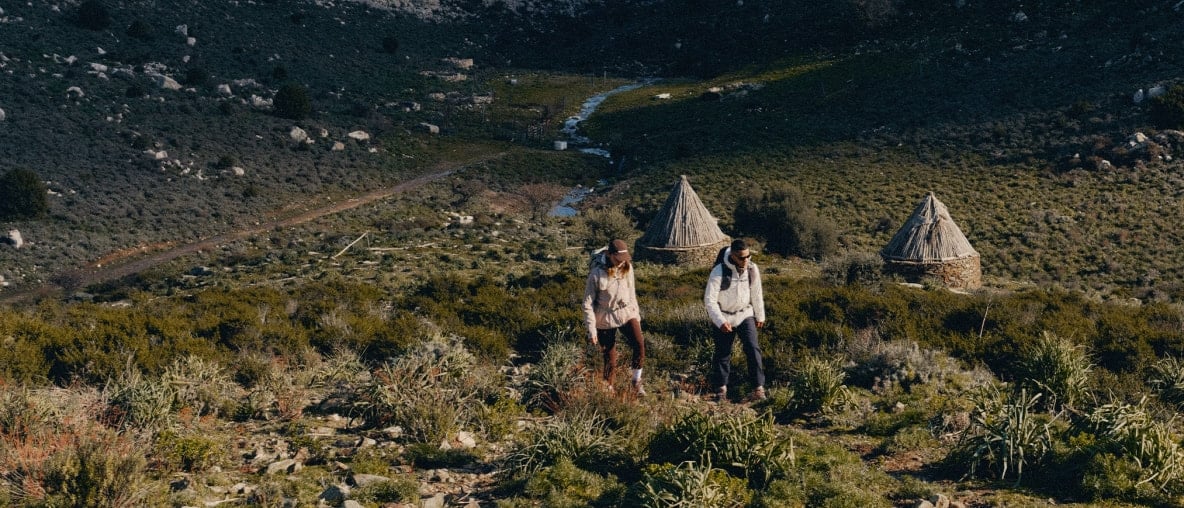 Zwei Wanderer in einer Berglandschaft mit traditionellen, runden Steinhütten.