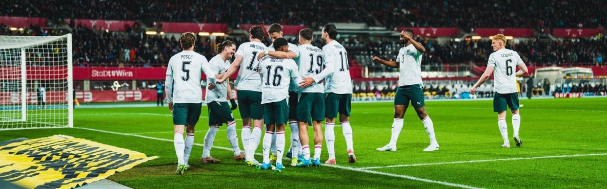 Mehrere Fußballspieler stehen dicht beieinander und jubeln auf dem Spielfeld nahe einem Tor. Im Hintergrund sind Tribünen mit Zuschauern in einem Stadion zu sehen.