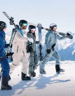 Vier Personen in Skibekleidung stehen im Schnee und tragen ihre Ski auf der Schulter mit Bergpanorama im Hintergrund.