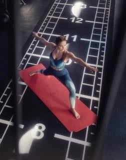 Frau steht auf einer roten Matte in einer ausbalancierten Yoga-Position mit ausgestreckten Armen. Die Übung fördert Gleichgewicht und Körperstabilität.