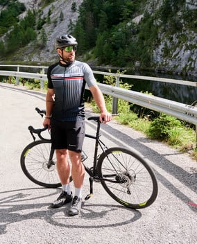 Mann steht neben einem Rennrad an einer Straße mit Geländer vor einem Fluss und Felsen.