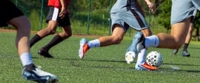 Mehrere Fußballspieler laufen auf einem Kunstrasenplatz einem Ball nach. Sichtbar sind Beine, Fußballschuhe und Trainingskleidung während einer Spielsituation.