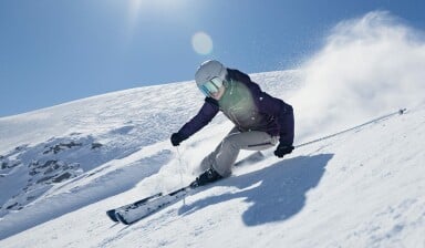 Eine Skifahrerin carvt bei Sonnenschein eine schneebedeckte Piste hinunter.