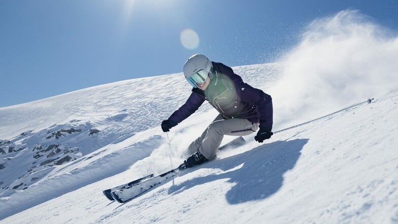 Eine Skifahrerin carvt bei Sonnenschein eine schneebedeckte Piste hinunter.
