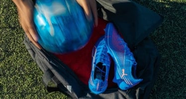 Offene Sporttasche auf Kunstrasen mit blauen Fußballschuhen, während ein Ball hineingelegt wird. Der Fokus liegt auf Ausrüstung und Tasche von oben.