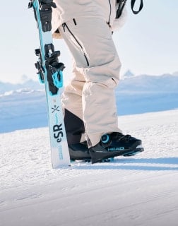 Nahaufnahme von Beinen in Skischuhen und einer Hand, die ein Paar Ski auf festem Schnee hält.