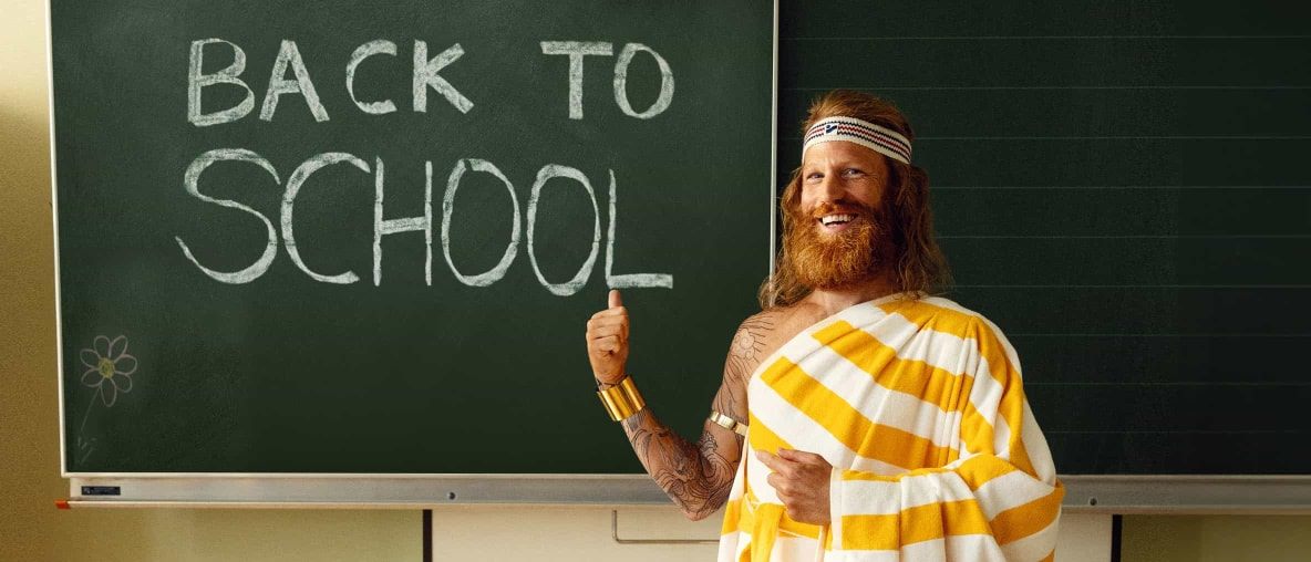Der Sommer steht vor einer dunkelgrünen Schultafel, auf der mit weißer Kreide "Back To School" geschrieben steht und lächelt fröhlich.