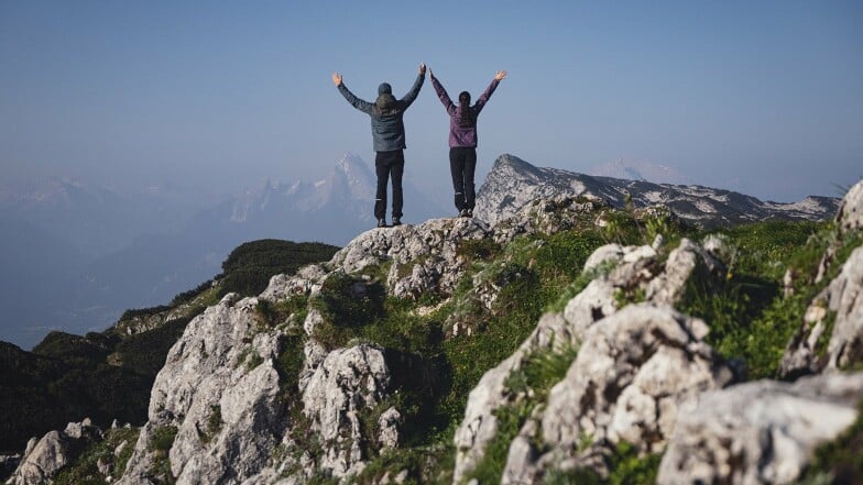 Zwei Wanderer jubeln auf einem Berggipfel mit herrlicher Aussicht.