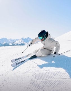 Eine Person in heller Skibekleidung fährt eine verschneite Piste hinunter und legt sich in die Kurve.