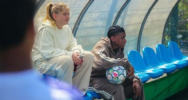 Zwei Menschen sitzen mit legerer Trainingskleidung und einem Ball auf der Ersatzbank eines Fußballfelds und beobachten das Spiel.