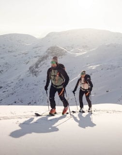 Zwei Personen mit Tourenski steigen bei Sonnenschein einen verschneiten Hang hinauf.