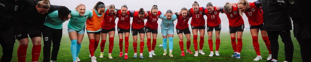 Mehrere Fußballspielerinnen stehen eng nebeneinander mit eingehakten Armen auf dem Spielfeld. Sie beugen sich nach vorne und rufen gemeinsam.