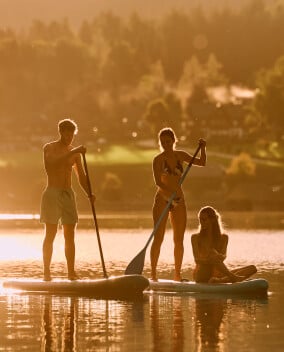 Drei Personen beim Stand-Up-Paddling auf einem ruhigen See im goldenen Gegenlicht der Abendsonne vor bewaldeten Bergen