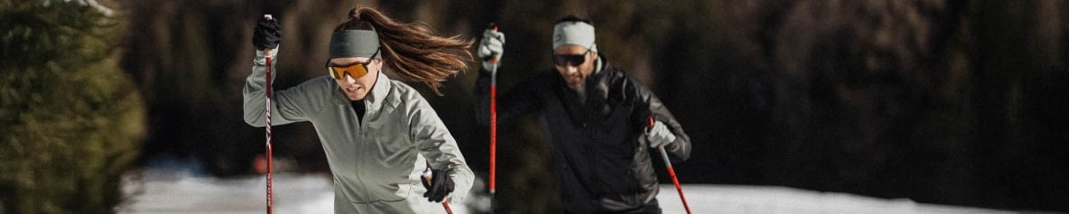 Zwei Personen betreiben Langlauf auf einer verschneiten Strecke im Wald in Odlo Langlaufbekleidung.