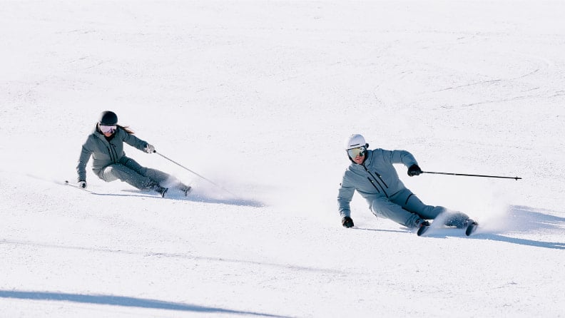 Zwei Skifahrer in grauen Anzügen carven auf einer schneebedeckten Piste.