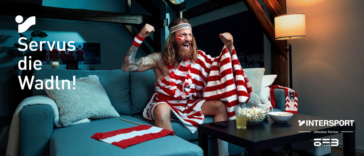 Jubelnder österreichischer Fußballfan mit rot-weißem Fan-Outfit feuert die Nationalmannschaft auf dem Sofa an.