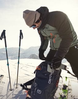 Eine Person in Winterkleidung packt im Schnee einen Rucksack neben Skistöcken und Skiern.