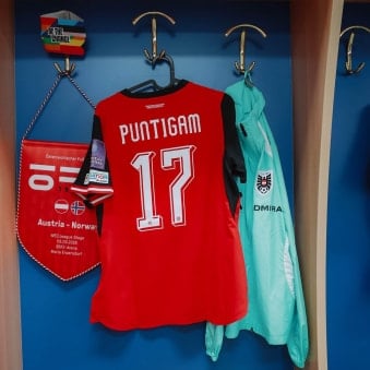 Ein rotes Fußballtrikot mit der Nummer 17 hängt an einem Haken neben einer Jacke. Daneben ist ein Wimpel an der Wand befestigt.