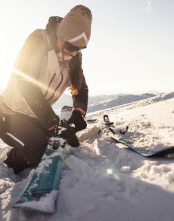Eine Person kniet im Schnee und befestigt die Bindung an einem Ski.
