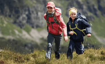 Dívka a chlapec jdou v přírodě s horami v pozadí oblečeni v outdoorovém oblečení.