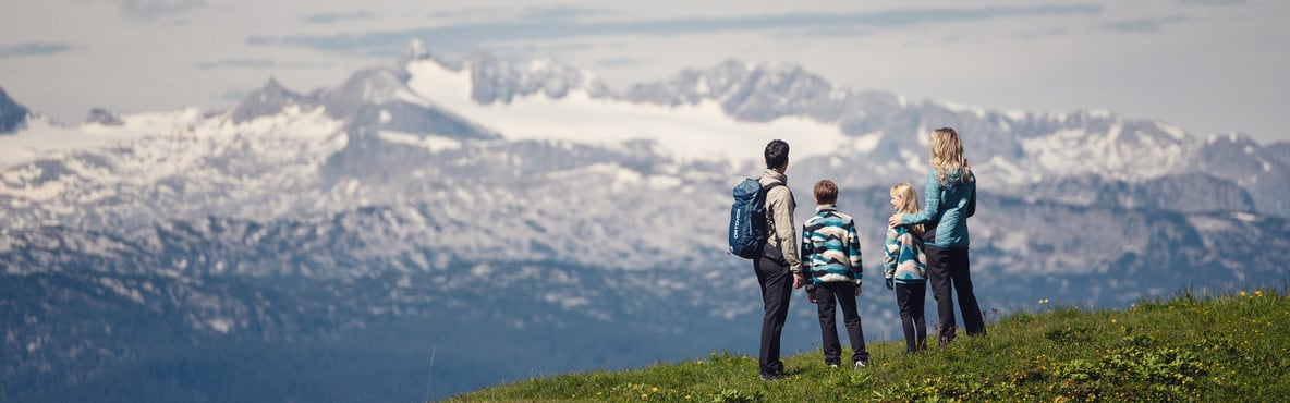 Vierköpfige Familie steht auf einer Bergwiese und blickt auf eine verschneite Berglandschaft