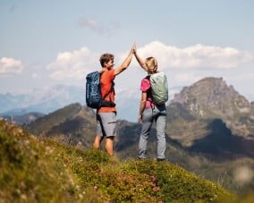 Zwei Wanderer mit Rucksäcken geben sich auf einem Berggipfel ein High Five vor einer Berglandschaft.