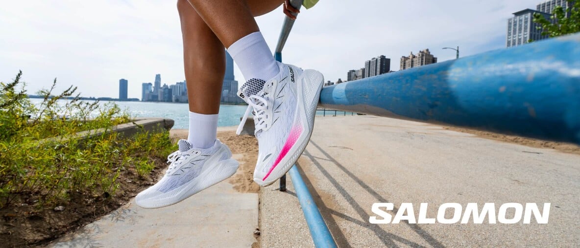 Beine mit weißen Laufschuhen ruhen auf einem Geländer am Wasser mit Stadtsilhouette im Hintergrund.