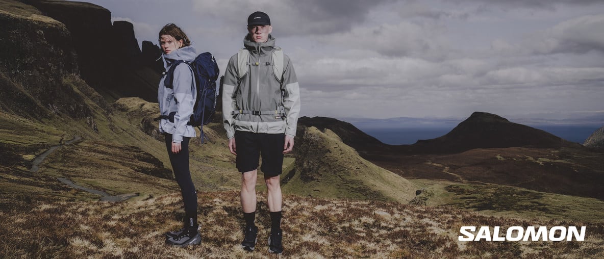 Zwei Personen in Outdoorbekleidung mit Rucksäcken vor bergiger Küstenlandschaft mit Salomon-Logo